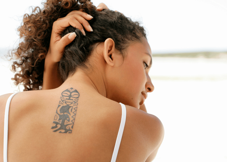 Young woman with curly hair showing a fresh black tattoo on her upper back, wearing a white strap top.